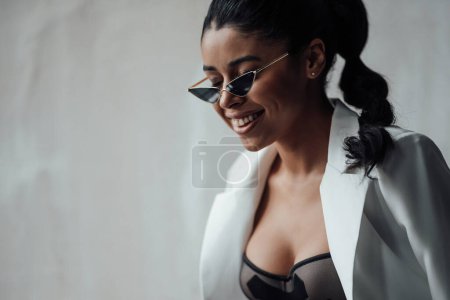 Photo for Young adult beauty swarthy woman in formal evening white suit with bra at cat eye sunglasses. Stylish black curly hair sensual african american model fashionista posing at studio in fashion pantsuit - Royalty Free Image