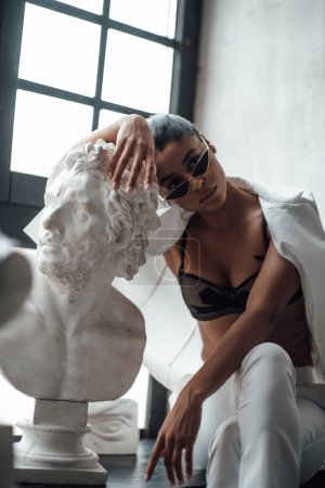 Photo for Young beauty swarthy woman in formal evening white suit no shirt hugs greek man bust statue. Stylish black curly hair sensual african american model fashionista posing at studio in fashion pantsuit - Royalty Free Image