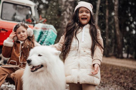 Photo for Little asian and caucasian girls spending time with white Samoyed dog near Xmas bus outdoor - Royalty Free Image