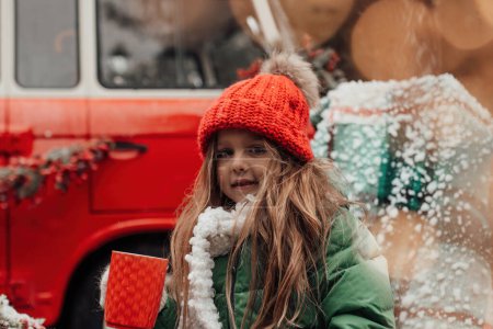 Téléchargez les photos : Enfant féminin célébrant Noël et Nouvel An saison des vacances d'hiver en plein air. Petite fille en tricot rouge chapeau joyeux passer du temps en plein air boire du chocolat chaud de grande tasse près de bus de Noël - en image libre de droit