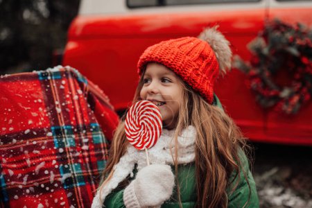 Foto de Niña celebrando Navidad y Año Nuevo vacaciones de invierno temporada al aire libre. Chica activa en rojo sombrero de punto alegre pasar tiempo al aire libre con caña de caramelo de Navidad en las manos - Imagen libre de derechos