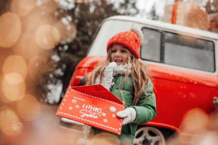 Photo for Female child celebrating Christmas and New Year winter holidays season outdoor. Little girl joyful spending time on open air holding letter to Santa in red envelope and waiting for miracle - Royalty Free Image