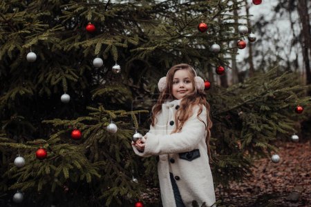 Téléchargez les photos : Petite fille active se reposer dans la forêt en appréciant les boules de décoration d'enfance arbre de Noël. Enfant féminin célébrant Noël et Nouvel An saison des vacances d'hiver en plein air - en image libre de droit