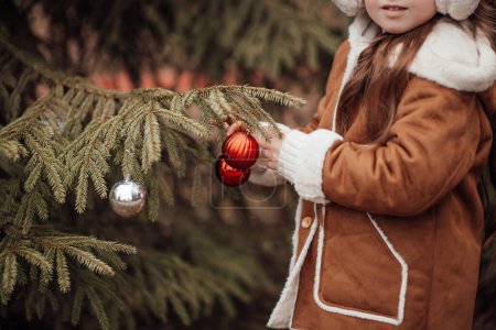 Téléchargez les photos : Petite fille active se reposer dans la forêt en appréciant les boules de décoration d'enfance arbre de Noël. Enfant féminin célébrant Noël et Nouvel An saison des vacances d'hiver en plein air - en image libre de droit