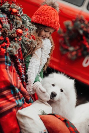 Foto de Niña celebrando Navidad y Año Nuevo vacaciones de invierno temporada al aire libre. Niña alegre pasar tiempo al aire libre con el perro Samoyedo blanco cerca de autobús de Navidad disfrutando de la infancia - Imagen libre de derechos
