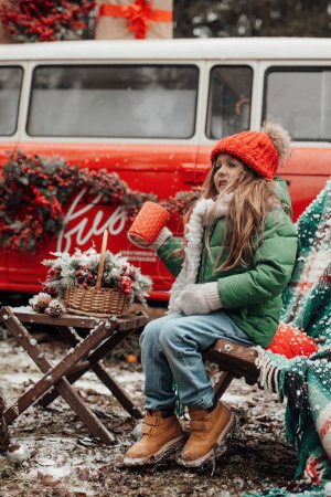 Photo for Female child celebrating Christmas and New Year winter holidays season outdoor. Little girl in red knitted hat joyful spending time on open air drinking hot chocolate from large mug near Xmas bus - Royalty Free Image