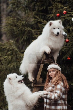 Foto de Niña celebrando Navidad y Año Nuevo vacaciones de invierno temporada al aire libre. Niña alegre pasar tiempo al aire libre con el perro Samoyedo blanco cerca de autobús de Navidad disfrutando de la infancia - Imagen libre de derechos