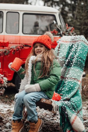 Téléchargez les photos : Enfant féminin célébrant Noël et Nouvel An saison des vacances d'hiver en plein air. Petite fille en tricot rouge chapeau joyeux passer du temps en plein air boire du chocolat chaud de grande tasse près de bus de Noël - en image libre de droit