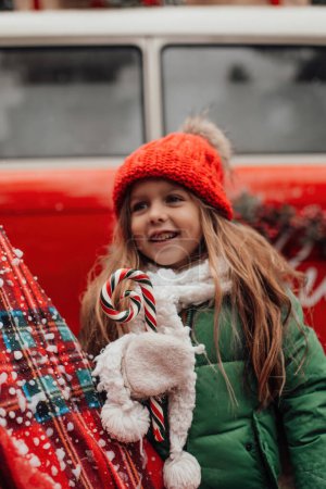 Foto de Niña celebrando Navidad y Año Nuevo vacaciones de invierno temporada al aire libre. Chica activa en rojo sombrero de punto alegre pasar tiempo al aire libre con caña de caramelo de Navidad en las manos - Imagen libre de derechos