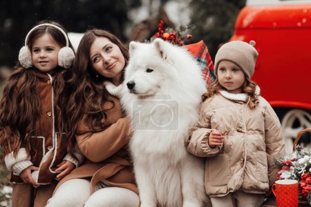 Photo for Happy family celebrating Christmas and New Year winter holidays season outdoor. Kids with mother and white Samoyed dogs joyful spending time together hugging near Xmas old bus - Royalty Free Image