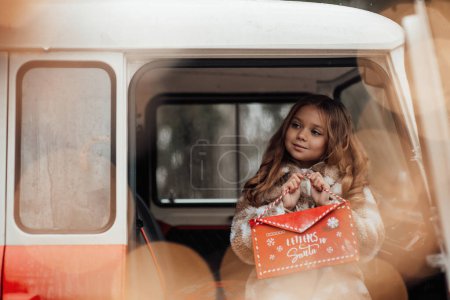 Photo for Female child celebrating Christmas and New Year winter holidays season outdoor. Little girl joyful spending time on open air holding letter to Santa in red envelope and waiting for miracle - Royalty Free Image