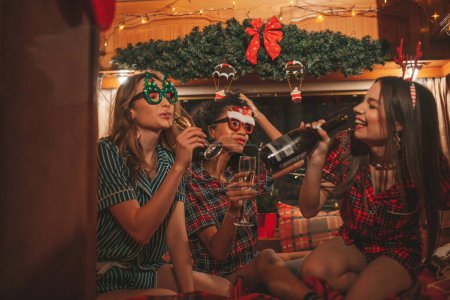 Téléchargez les photos : Filles heureuses célébrant Noël et Nouvel An saison des vacances d'hiver en camping-car. Actifs jeunes femmes diversifiées joyeux passer du temps ensemble étreignant s'amuser à boire du vin mousseux dans une vieille bande-annonce de Noël - en image libre de droit