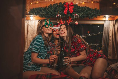 Foto de Chicas felices celebrando Navidad y Año Nuevo temporada de vacaciones de invierno en campista. Jóvenes activos diversas mujeres alegre pasar tiempo juntos abrazos divertirse beber vino espumoso en el viejo tráiler de Navidad - Imagen libre de derechos