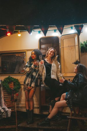 Foto de Chicas felices celebrando Navidad y Año Nuevo vacaciones de invierno temporada al aire libre. Jóvenes diversas mujeres alegres amigas pasando tiempo juntas se divierten bebiendo vino espumoso cerca del viejo tráiler de Navidad - Imagen libre de derechos