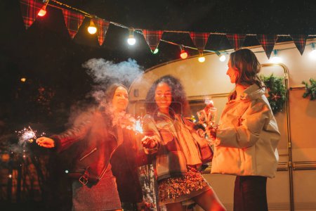 Foto de Chicas felices celebrando Navidad y Año Nuevo vacaciones de invierno temporada al aire libre. Jóvenes activos diversas mujeres alegres amigas junto con las luces de Bengala chispas se divierten cerca del viejo tráiler de Navidad - Imagen libre de derechos