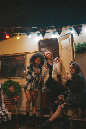 Foto de Chicas felices celebrando Navidad y Año Nuevo vacaciones de invierno temporada al aire libre. Jóvenes diversas mujeres alegres amigas pasando tiempo juntas se divierten bebiendo vino espumoso cerca del viejo tráiler de Navidad - Imagen libre de derechos