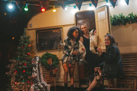 Photo for Happy girls celebrating Christmas and New Year winter holidays season outdoor. Young diverse women joyful female friends spending time together have fun drinking sparkling wine near old xmas trailer - Royalty Free Image