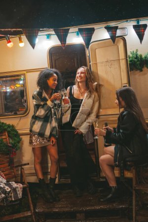 Photo for Happy girls celebrating Christmas and New Year winter holidays season outdoor. Young diverse women joyful female friends spending time together have fun drinking sparkling wine near old xmas trailer - Royalty Free Image
