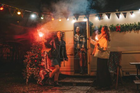 Photo for Happy girls celebrating Christmas and New Year winter holidays season outdoor. Active young diverse women joyful female friends together with Bengal lights sparklers have fun near old Xmas trailer - Royalty Free Image