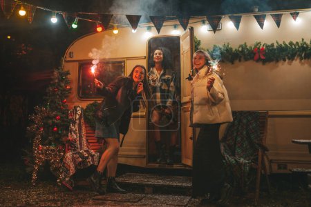 Téléchargez les photos : Filles heureuses célébrant Noël et Nouvel An saison des vacances d'hiver en plein air. Jeunes femmes actives diverses joyeuses amies avec des lumières du Bengale scintillantes s'amusent près de vieux trailer de Noël - en image libre de droit