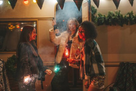 Photo for Happy girls celebrating Christmas and New Year winter holidays season outdoor. Active young diverse women joyful female friends together with Bengal lights sparklers have fun near old Xmas trailer - Royalty Free Image