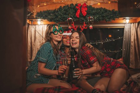 Téléchargez les photos : Filles heureuses célébrant Noël et Nouvel An saison des vacances d'hiver en camping-car. Actifs jeunes femmes diversifiées joyeux passer du temps ensemble étreignant s'amuser à boire du vin mousseux dans une vieille bande-annonce de Noël - en image libre de droit