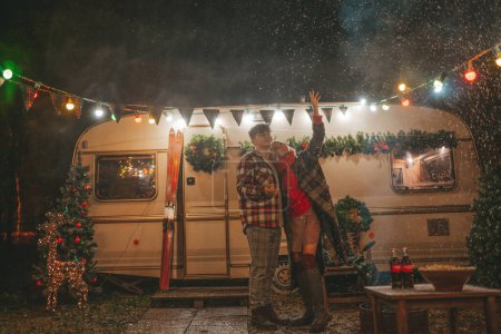 Téléchargez les photos : Joyeux couple célébrant Noël et Nouvel An saison des vacances d'hiver dans Camper Park. Jeune couple joyeux passer du temps ensemble câlins et bisous se réjouit à la première neige près de caravane de camping-car de Noël - en image libre de droit