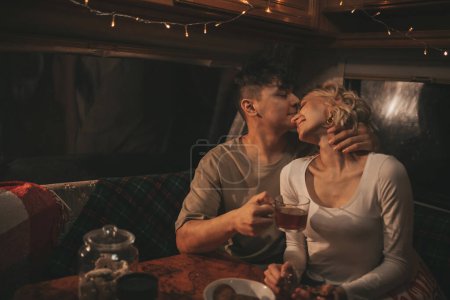 Photo for Happy couple celebrating Christmas and New Year winter holidays season in camper. Young couple drinking tea spending time together hugs and kisses in Xmas camper trailer - Royalty Free Image