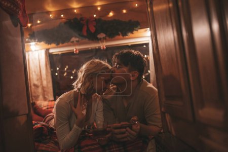 Téléchargez les photos : Joyeux couple célébrant Noël et Nouvel An saison des vacances d'hiver en camping-car. Jeune couple boire du thé passer du temps ensemble câlins et bisous dans la caravane de Noël - en image libre de droit