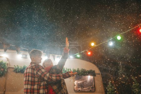 Photo for Happy couple celebrating Christmas and New Year winter holidays season in Camper Park. Young joyful couple spending time together hugs and kisses rejoices at first snow near Xmas camper trailer - Royalty Free Image