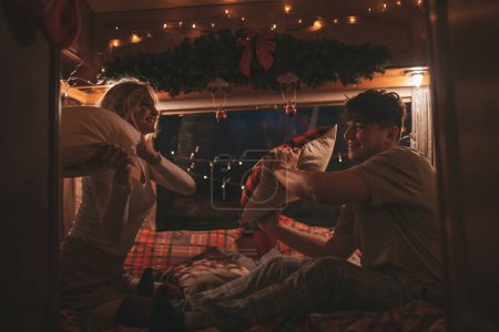 Téléchargez les photos : Joyeux couple célébrant Noël et Nouvel An saison des vacances d'hiver en camping-car. Jeune couple joyeux passer du temps ensemble avoir plaisir bataille d'oreillers dans la caravane de camping-car de Noël - en image libre de droit