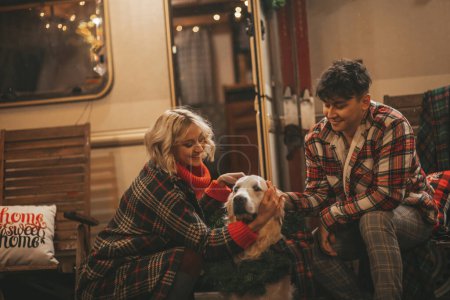 Photo for Happy couple celebrating Christmas and New Year winter holidays season in Camper Park. Young joyful couple hugging and spending time together with golden retriever dog near Xmas camper trailer - Royalty Free Image