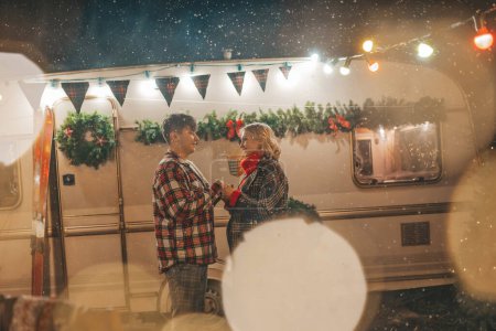 Téléchargez les photos : Joyeux couple célébrant Noël et Nouvel An saison des vacances d'hiver dans Camper Park. Jeune couple joyeux passer du temps ensemble câlins et bisous se réjouit à la première neige près de caravane de camping-car de Noël - en image libre de droit