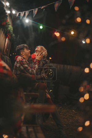 Photo for Happy couple celebrating Christmas and New Year winter holidays season in Camper Park. Young couple rest and relaxation spending time together hugs and kisses near Xmas camper trailer - Royalty Free Image