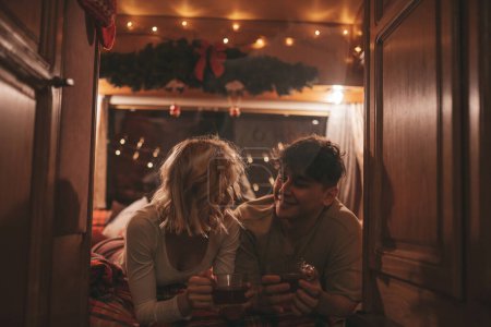 Téléchargez les photos : Joyeux couple célébrant Noël et Nouvel An saison des vacances d'hiver en camping-car. Jeune couple boire du thé passer du temps ensemble câlins et bisous dans la caravane de Noël - en image libre de droit