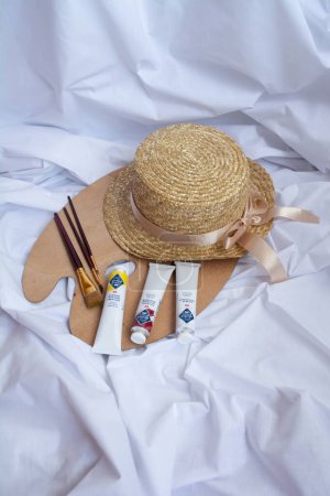 Photo for This photo captures the beauty of oil painting with its vibrant colors, texture, and artistry. The photo features a straw hat, an artistic wooden paint palette, and various tubes of oil paint, all laid out on a clean, white canvas. - Royalty Free Image