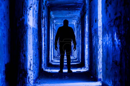 Sylwetka mężczyzny w długim ciemnym, przerażającym korytarzu, fioletowo-niebieska wersja stonowana. Koncepcja horroru lub zombie