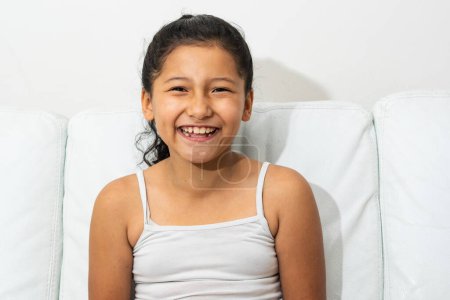 Porträt einer lateinischen Brünetten, die lächelnd auf dem Sofa sitzt und ihre Zähne zeigt, Mädchen mit indischen Zügen