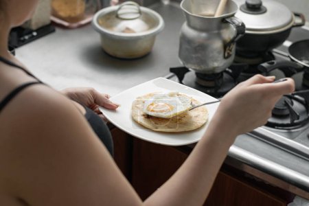 Foto de Mujer latina preparando un típico desayuno colombiano de arepa con huevo, chica en su cocina preparando comida - Imagen libre de derechos