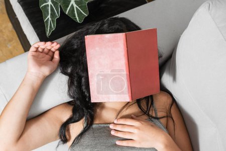Foto de Chica morena acostada en su sofá con un libro rojo colocado en su cara. mujer latina dormida leyendo un libro. - Imagen libre de derechos