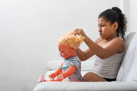 chica morena muy concentrada jugando con su muñeca sentada en el sofá de la sala de estar, chica latina jugando a ser una mamá.