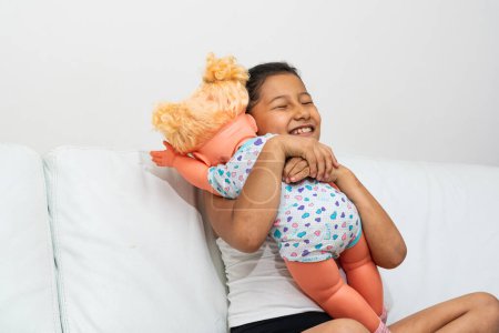 chica morena latina, muy feliz abrazando a su muñeca mientras juega que ella es su hija. la chica está emocionada con su nuevo juguete.