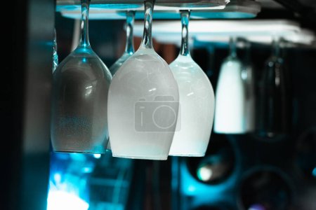 Photo for White wine glasses hanging over the top, in a liquor cabinet in a bar, with an ambiance of blue light on a black background. - Royalty Free Image