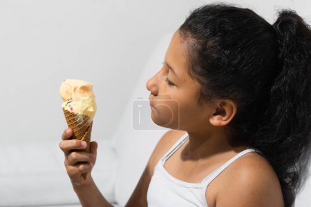 Téléchargez les photos : Little brown-skinned latina girl, holding an ice cream cone in her hand, looking at it before eating it - en image libre de droit