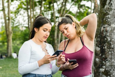 Photo for Latina women in the park, one with a look of discomfort at what she is looking at on her friend's cell phone. - Royalty Free Image