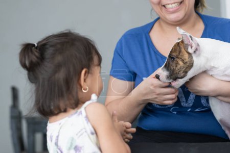 Téléchargez les photos : Latina mère jouer avec son taureau terrier chien alors qu'il mord elle et sa petite fille regarde - en image libre de droit