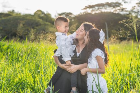 Téléchargez les photos : Famille latino, jeune mère avec ses enfants, tenant son jeune fils tout en souriant, avec un coucher de soleil en arrière-plan. - en image libre de droit