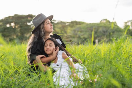 young latin mother with her daughter lying on her chest, sitting on the tall grass while the woman thanks the sky and asks god