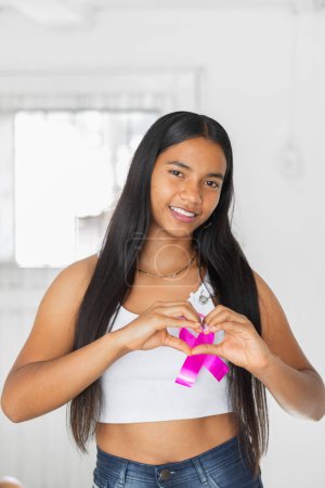 joven morena latina haciendo un corazón con las manos, con una cinta rosa símbolo de la lucha contra el cáncer de mama.
