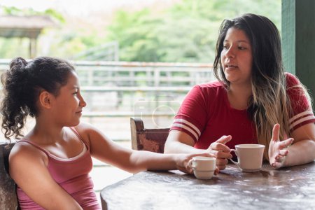 young latina mother talking to her daughter while drinking coffee at the dinner table